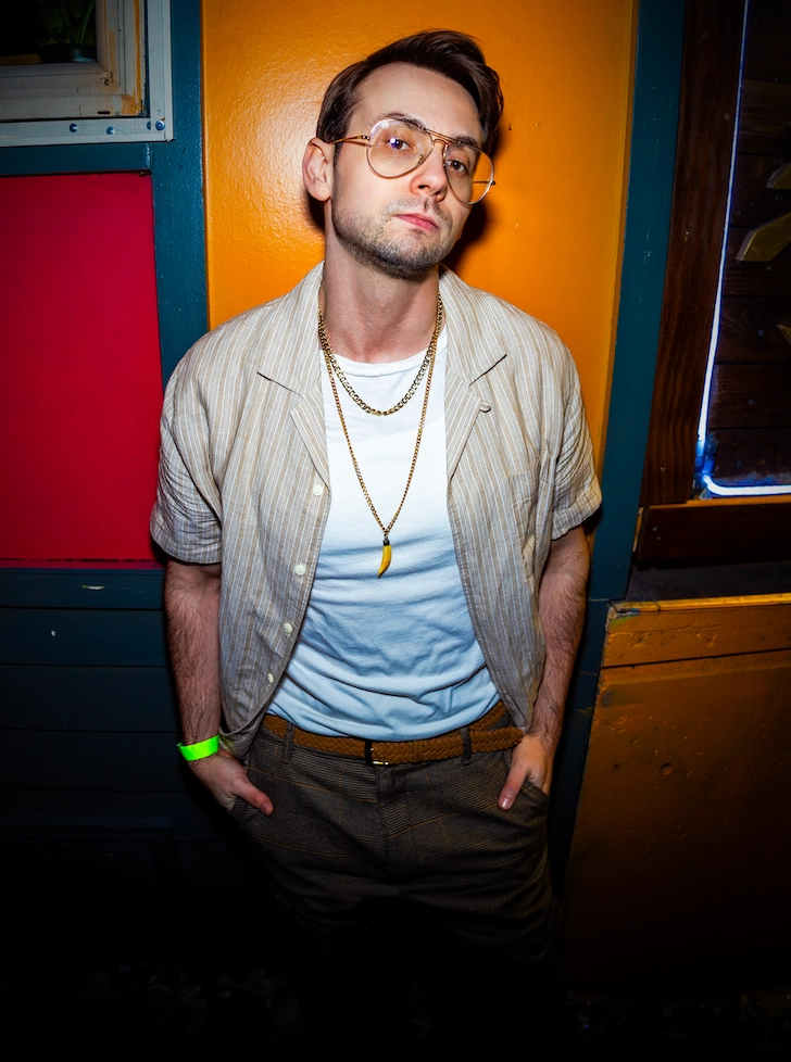 Portrait photo of Kellan leading against a wall wearing a white t-shirt, gold necklaces, and plaid pants outside of Nectar Lounge in Seattle.