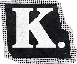 Shortened logo for Kellan just showing the "K" in bold, white, distressed capital letters on a black ticket-like background with dotted borders. The text and background have a textured, vintage appearance, and there's a period after the name.